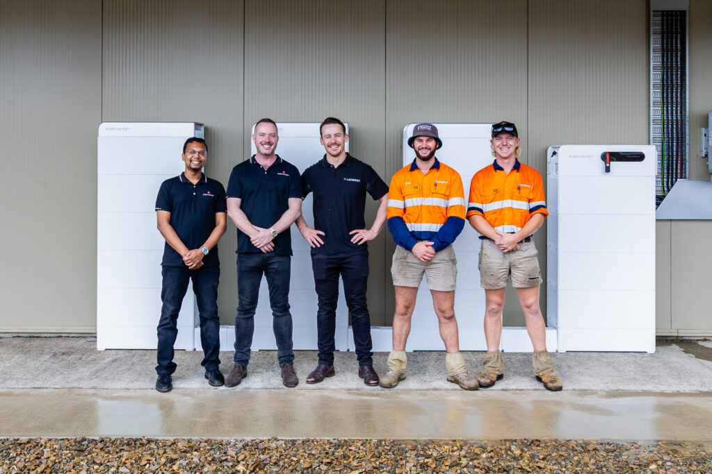 Five men standing in front of home Sigenergy Sigenstack battery systems, including Lenergy installers in safety gear and staff in branded shirts at installation site
