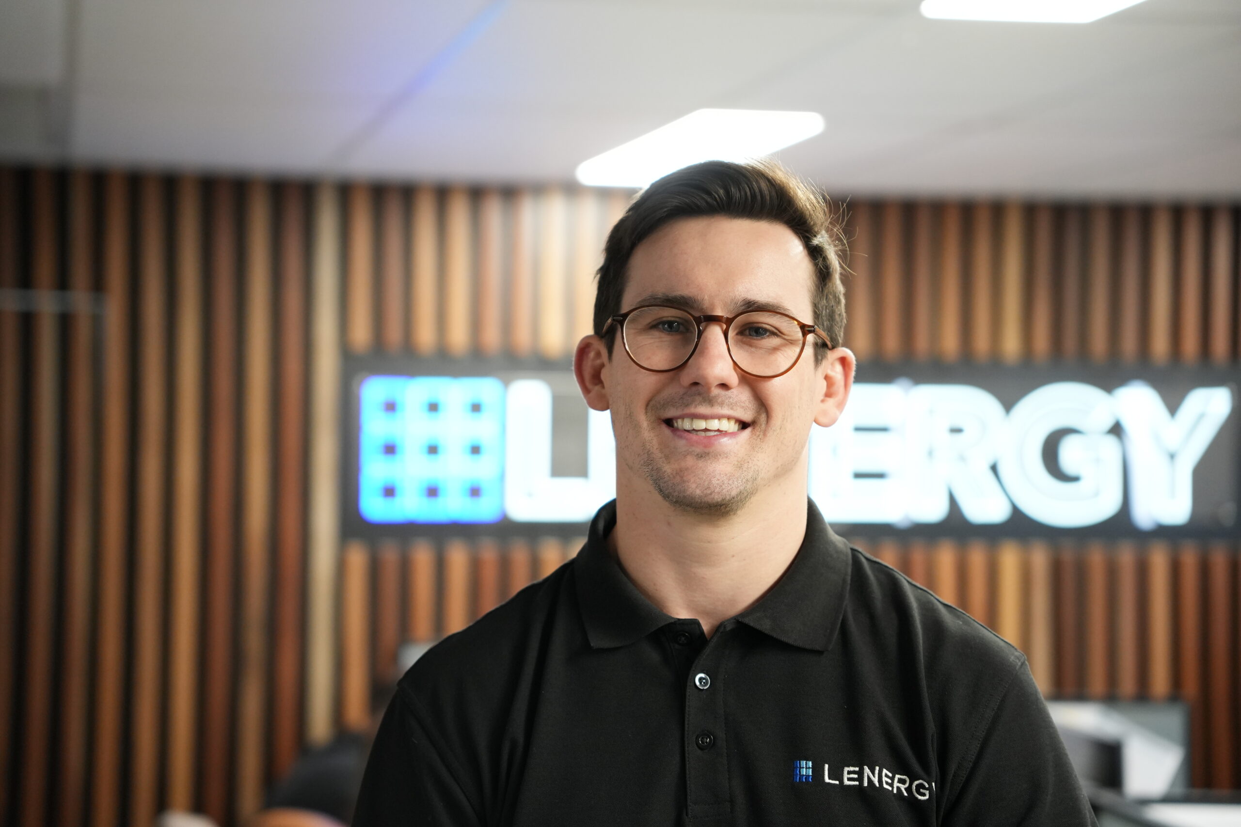 Smiling installer wearing glasses and black Lenergy polo shirt standing in office with company logo on wall behind him