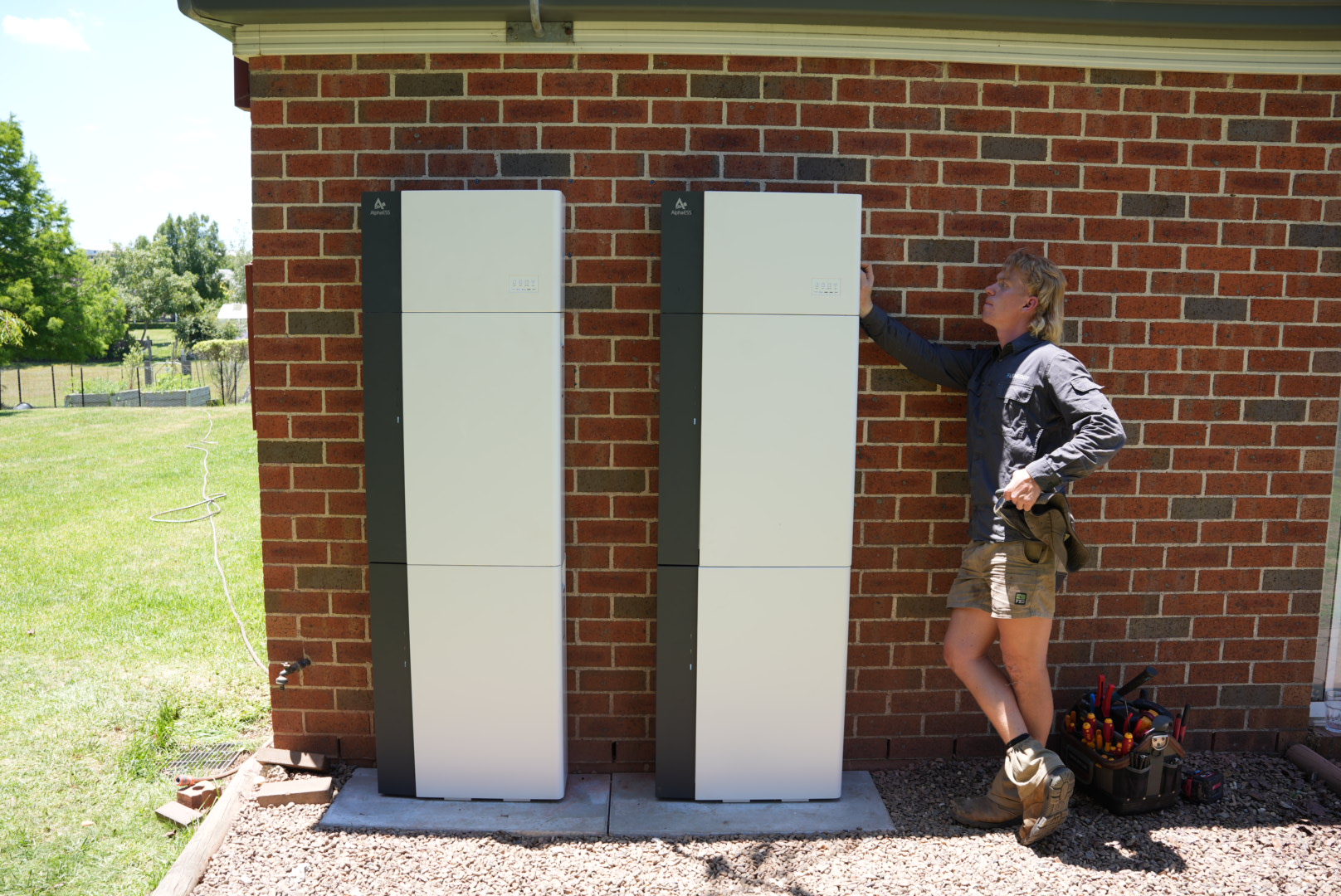 Technician inspecting two wall-mounted Alpha ESS Battery units on a brick house exterior, with tools nearby and grassy yard visible