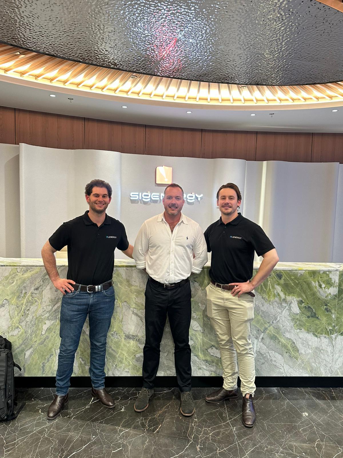 Three men standing together in a lobby at a conference venue, smiling in front of a marble reception desk and modern interior backdrop