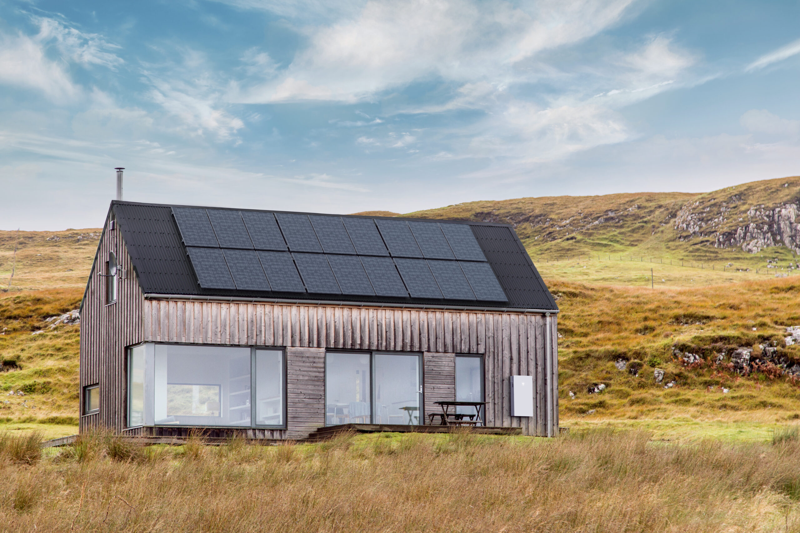 Modern rural home with rooftop solar panels and Tesla Powerwall 3 on exterior wall, set in grassy landscape under blue sky