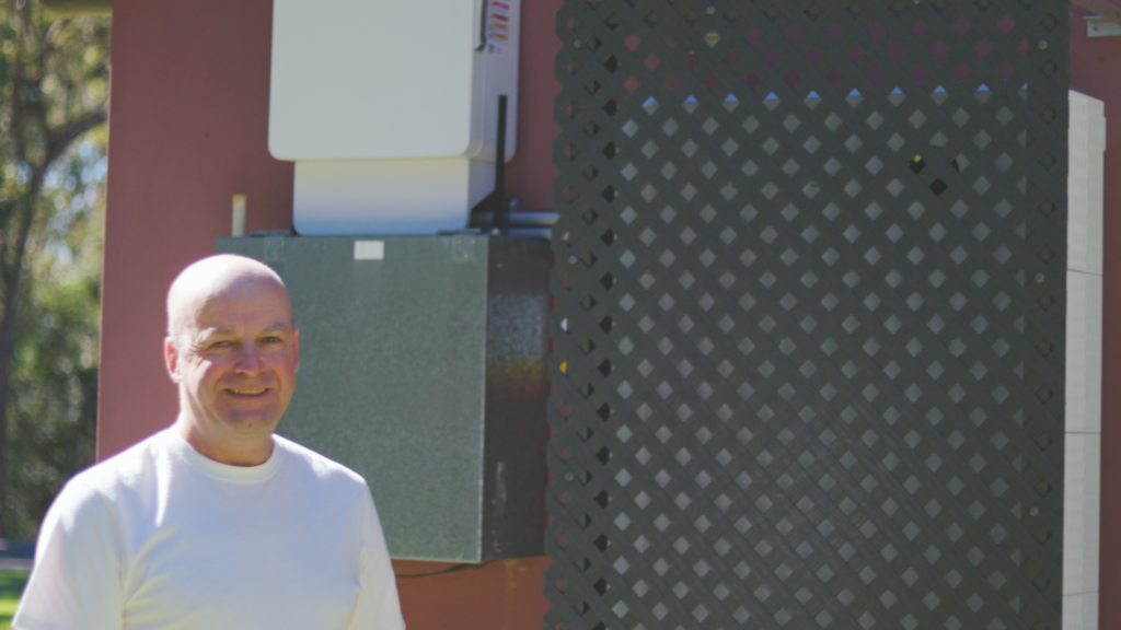 Peter stands beside a home solar battery and inverter, with the battery partly screened by a protective lattice outdoors.