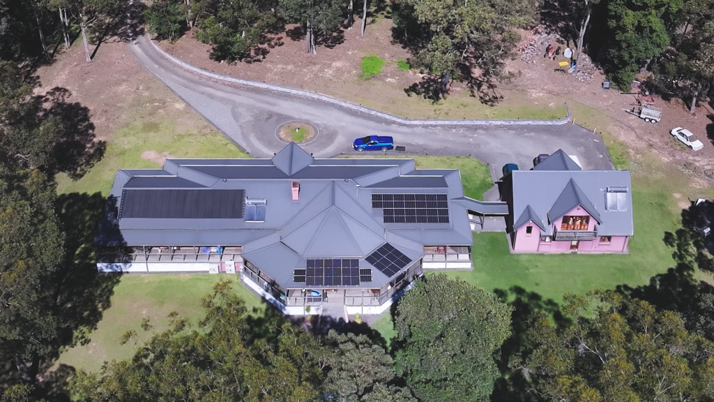 Aerial view of a large rural home with multiple rooftop solar panel arrays, surrounded by trees and open grounds on a sunny day.