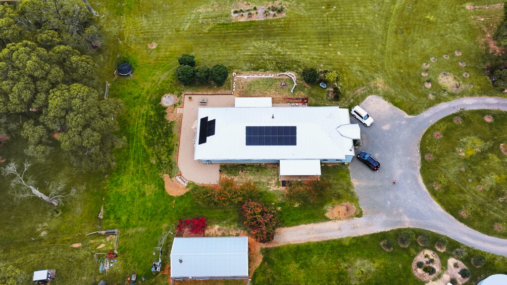 Aerial view of a rural home with rooftop solar panels, surrounded by gardens, trees and a long country driveway.
