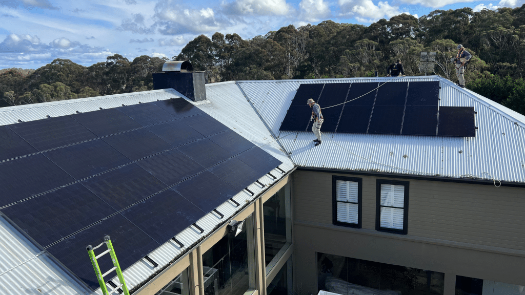 Two workers install solar panels on the metal roof of a large house, with multiple panel arrays already in place and a wooded landscape in the background under a partly cloudy sky.