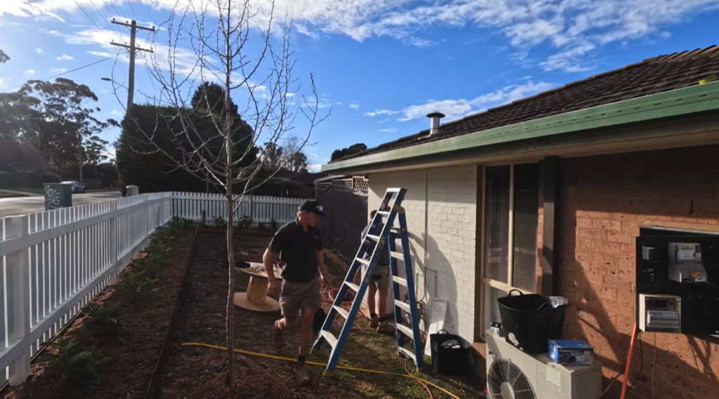 A Lenergy technician carrying equipment during a solar battery installation beside a brick home, with a ladder against the wall and clear blue sky overhead.