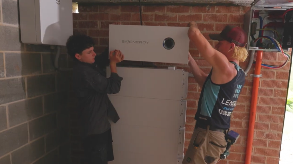 Two Lenergy installers mounting a stacked Sigenergy SigenStor battery system against the wall beside the switchboard