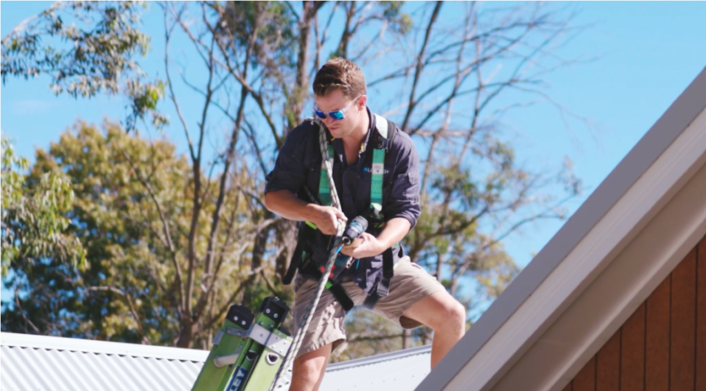 A young installer on a rooftop, wearing a safety harness and using a drill during a solar panel installation.