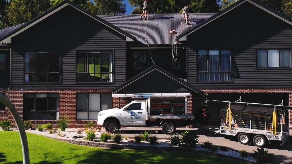 Lenergy installers harnessed on the roof of a two-storey Burradoo home, with a branded Lenergy ute and trailer in the driveway.