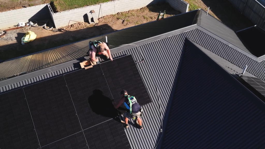 Aerial view of two harnessed Lenergy installers fitting black solar panels to a dark metal roof during a Burradoo solar install.