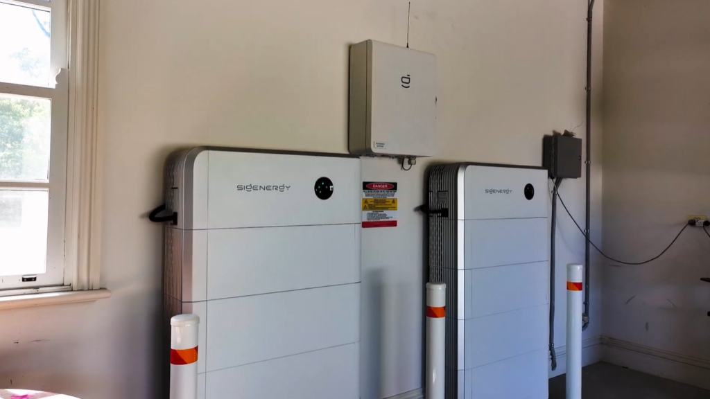 Two wall-mounted Sigenergy SigenStor battery units installed in a garage, with a controller box above and safety bollards in front