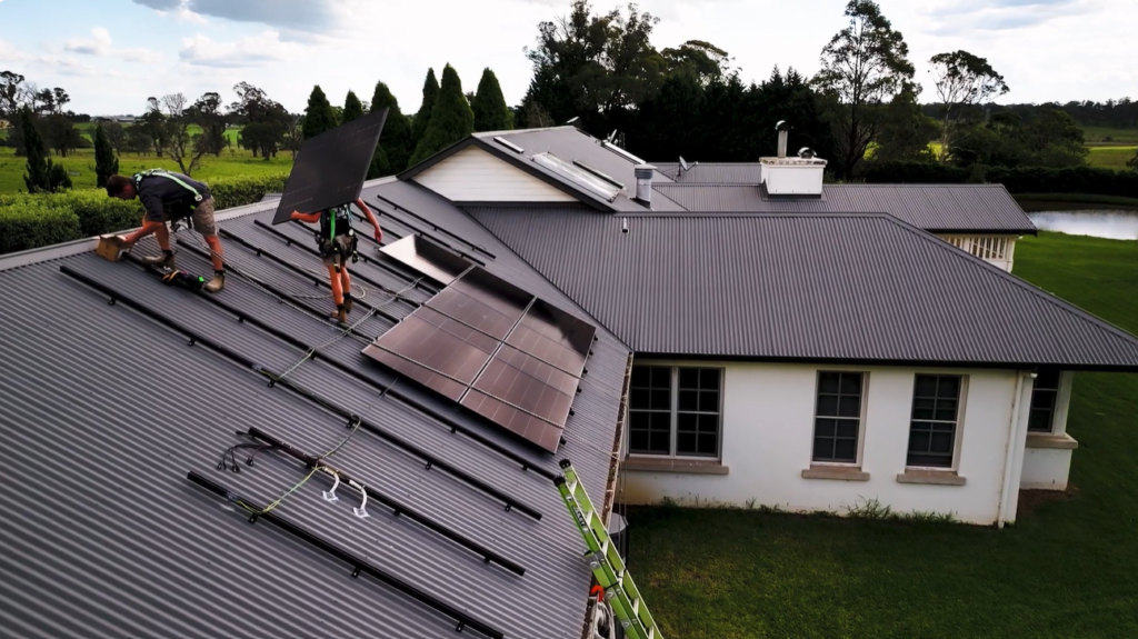 Two installers fitting solar panels onto the charcoal metal roof of a rural home, with mounting rails and part-completed rows in place