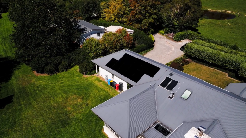 Aerial view of a large rural property with black solar panels mounted on an outbuilding roof, surrounded by lawn and trees