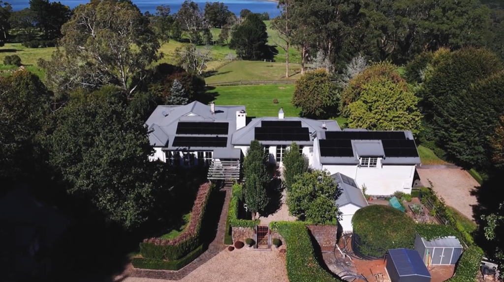 Aerial view of Mike Chapman's Southern Highlands home with rooftop solar panels, set among green lawns and trees with water in the distance.