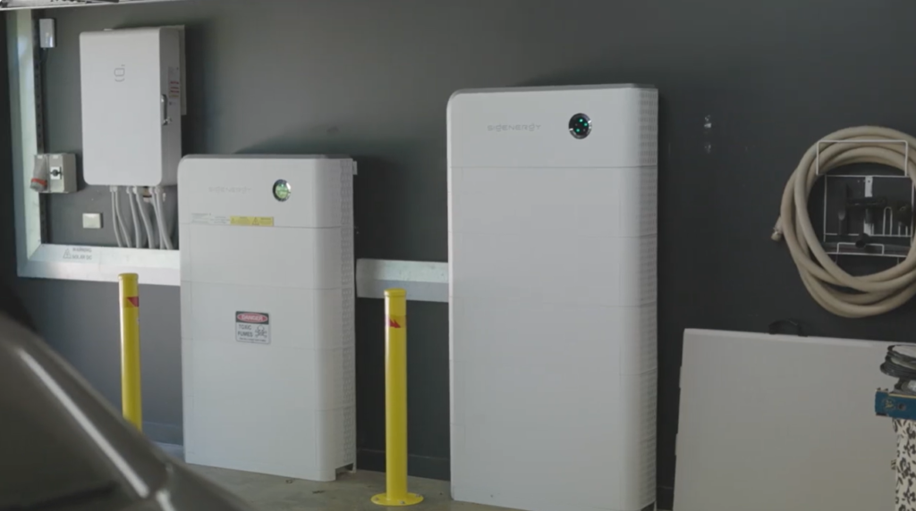 Mike's Sigenergy SigenStor battery system installed in his garage, two white units against a dark grey wall with a wall-mounted gateway and yellow safety bollards.